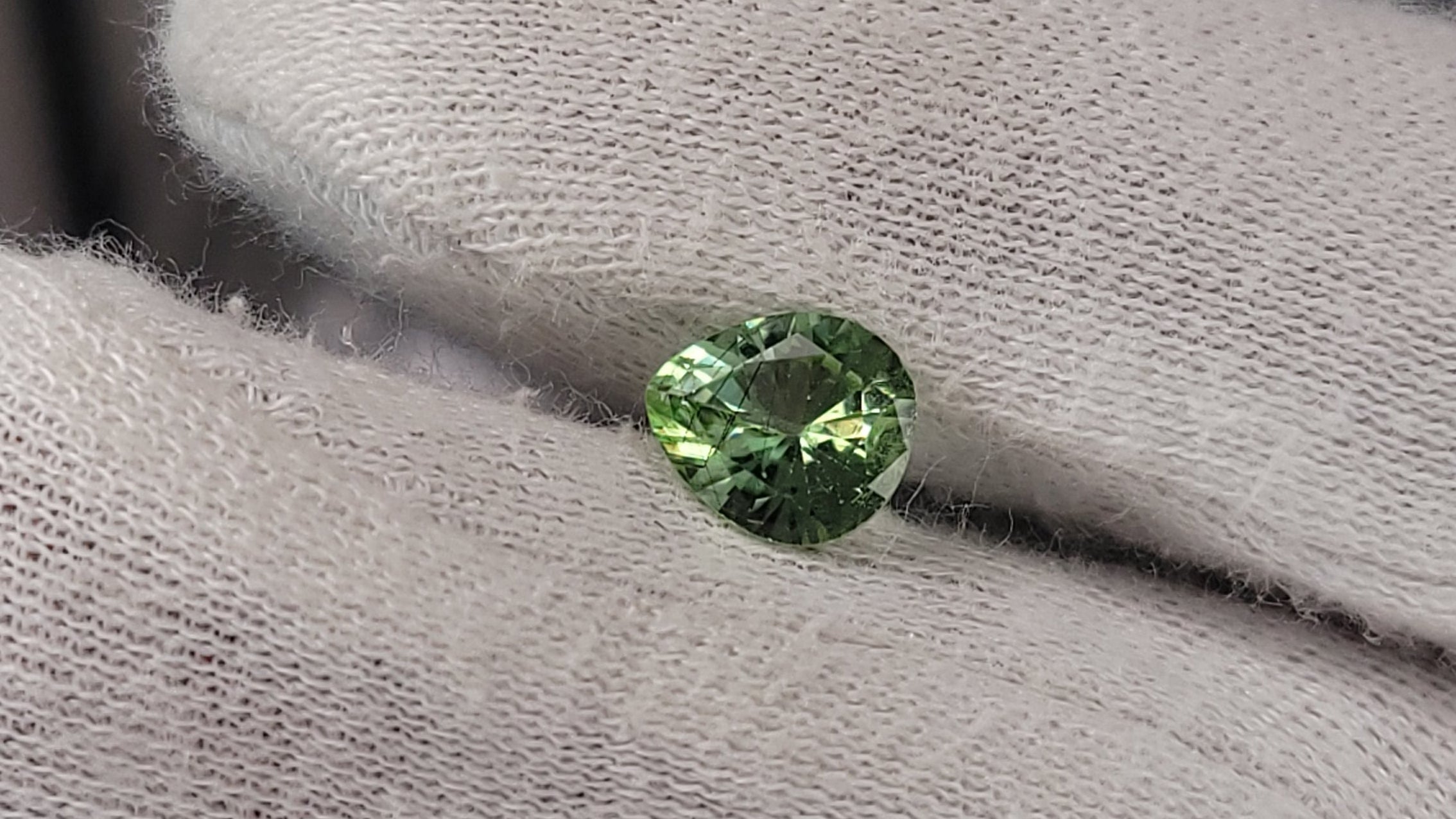 A small round green gemstone held between white gloved fingers.