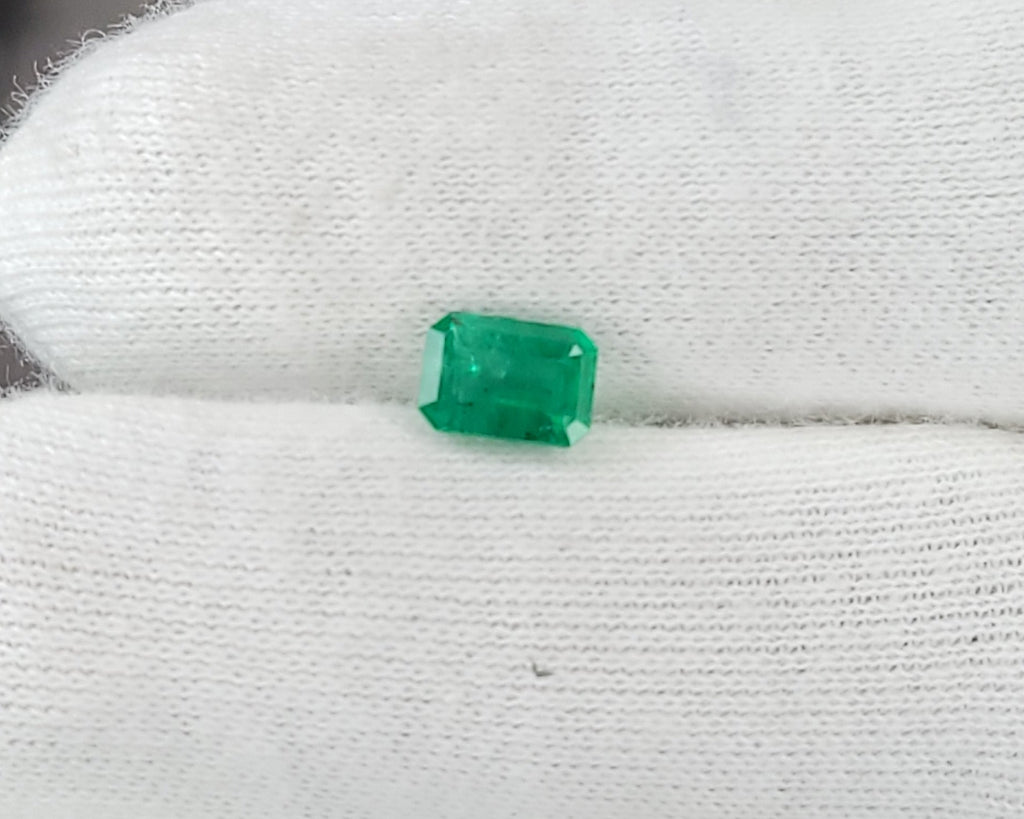 A small rectangular green gemstone held between white gloved fingers, with a blurred studio or workshop background.
