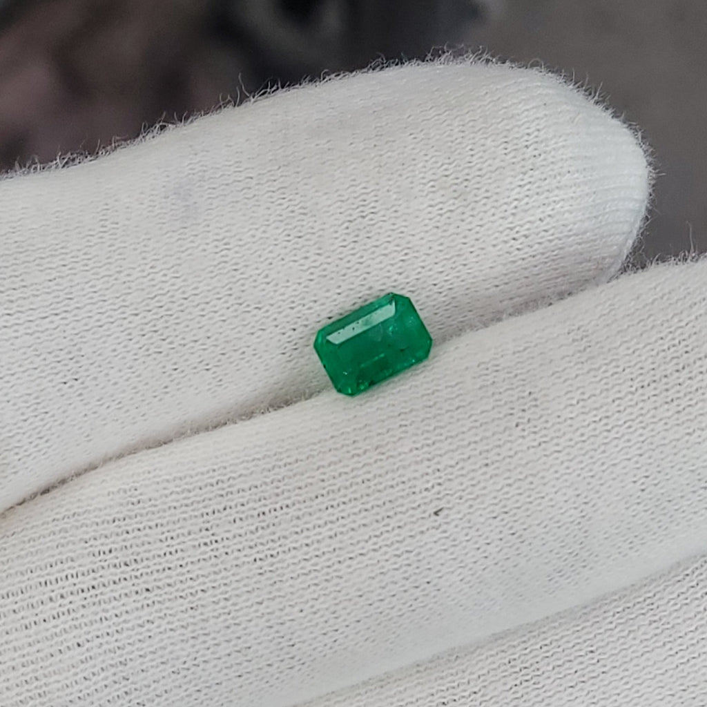 A small rectangular green gemstone held on a white knit-gloved finger, with a blurred industrial background.