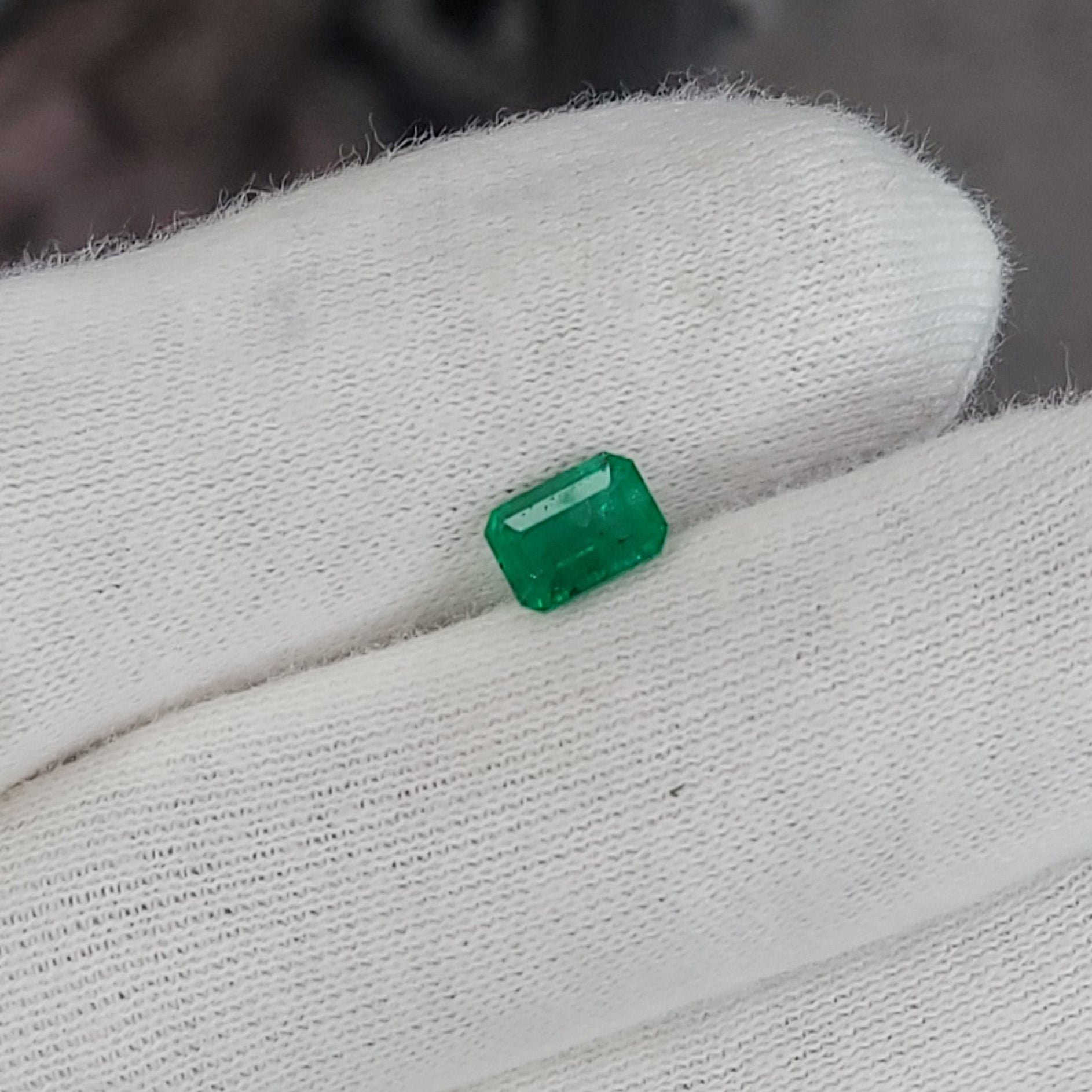 A small rectangular green gemstone held on a white knit-gloved finger, with a blurred industrial background.