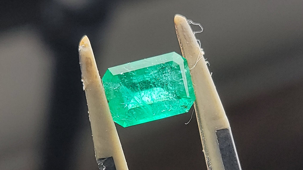 A green rectangular gemstone held between a pair of tweezers against a dark, blurred background.