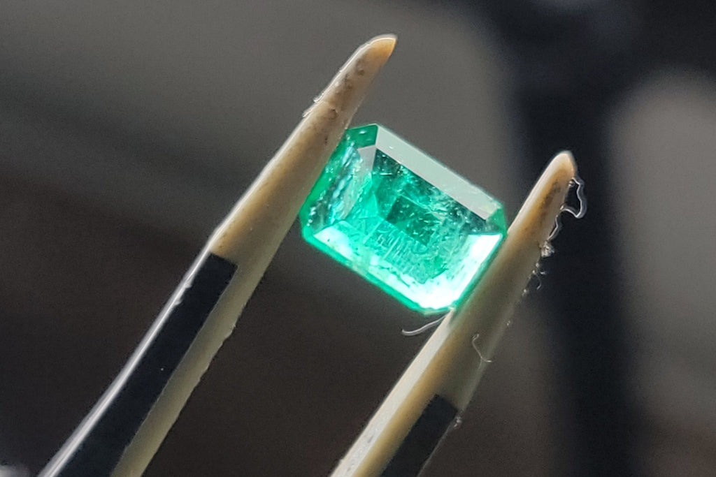 A rectangular green gemstone held between metal tweezers against a dark blurred background.