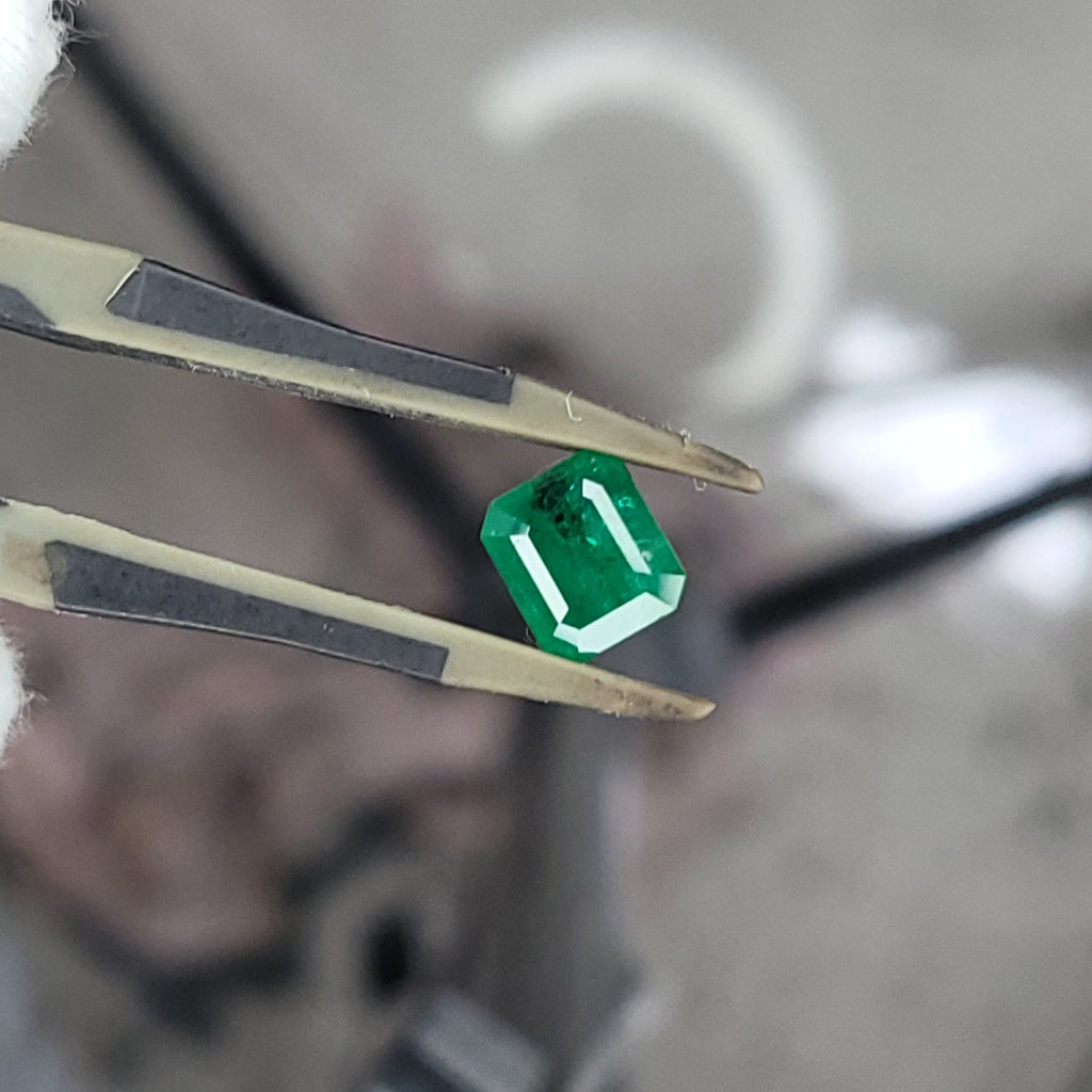A small green faceted gemstone held between metal tweezers, photographed close-up against a blurred workshop-like background.