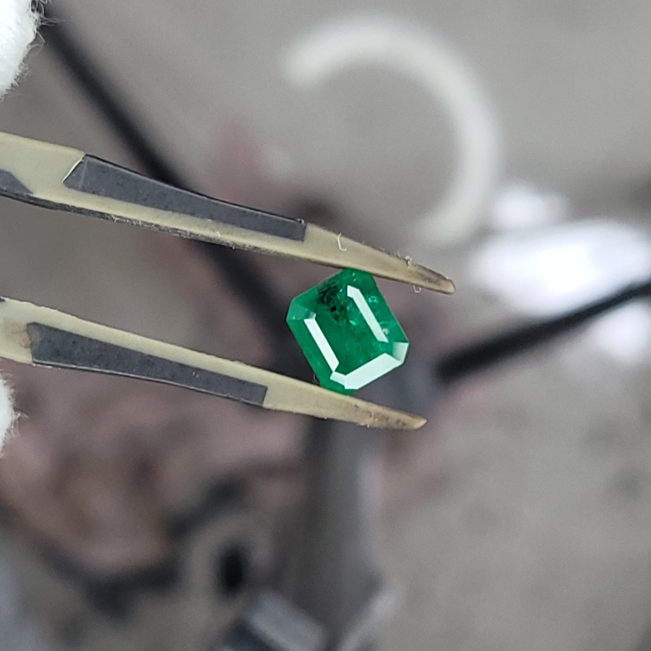 A small green faceted gemstone held between metal tweezers, photographed close-up against a blurred workshop-like background.