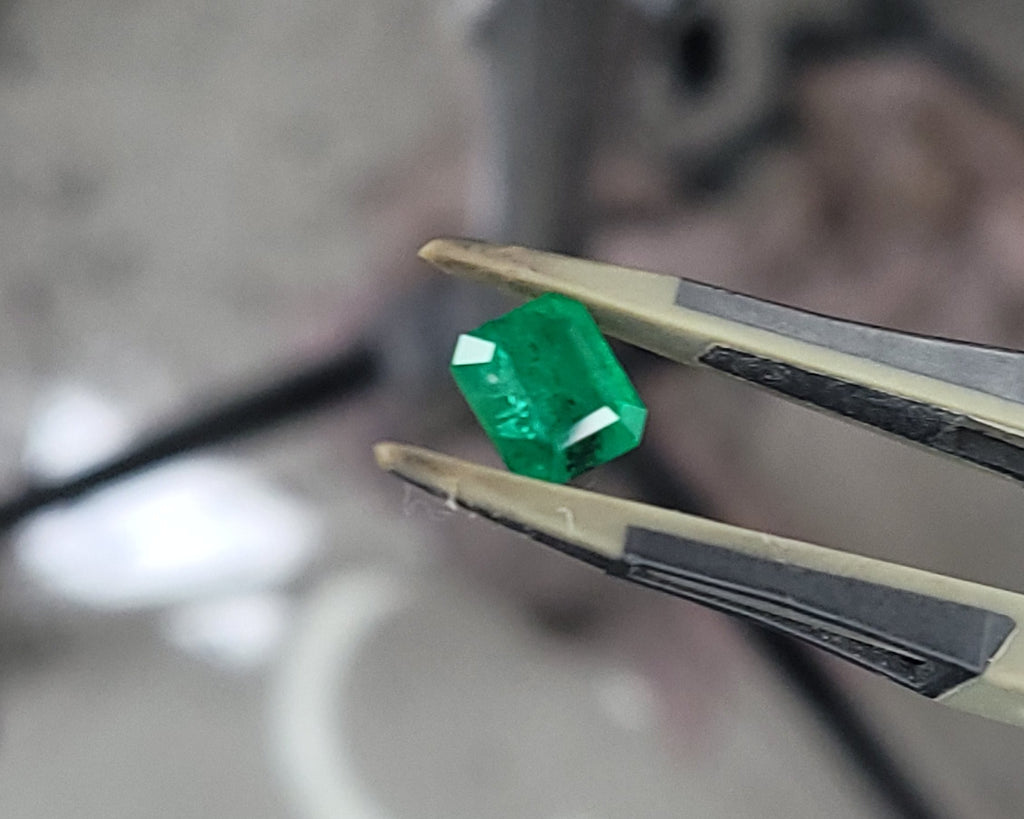A bright green emerald-cut gemstone held between metal tweezers, with a blurred workshop or studio background.