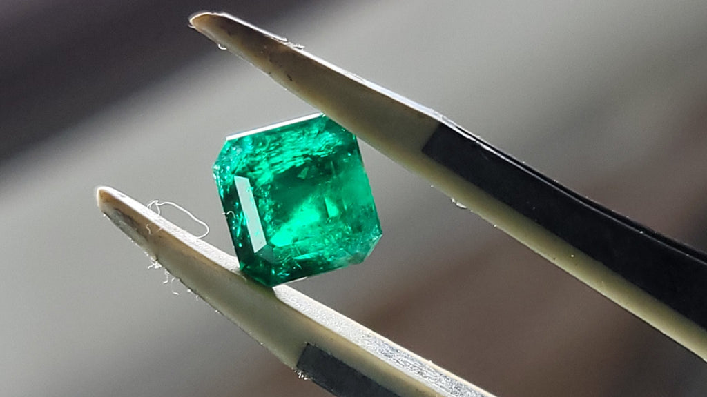 A green emerald-cut gemstone held between two metal tweezers against a blurred indoor background.