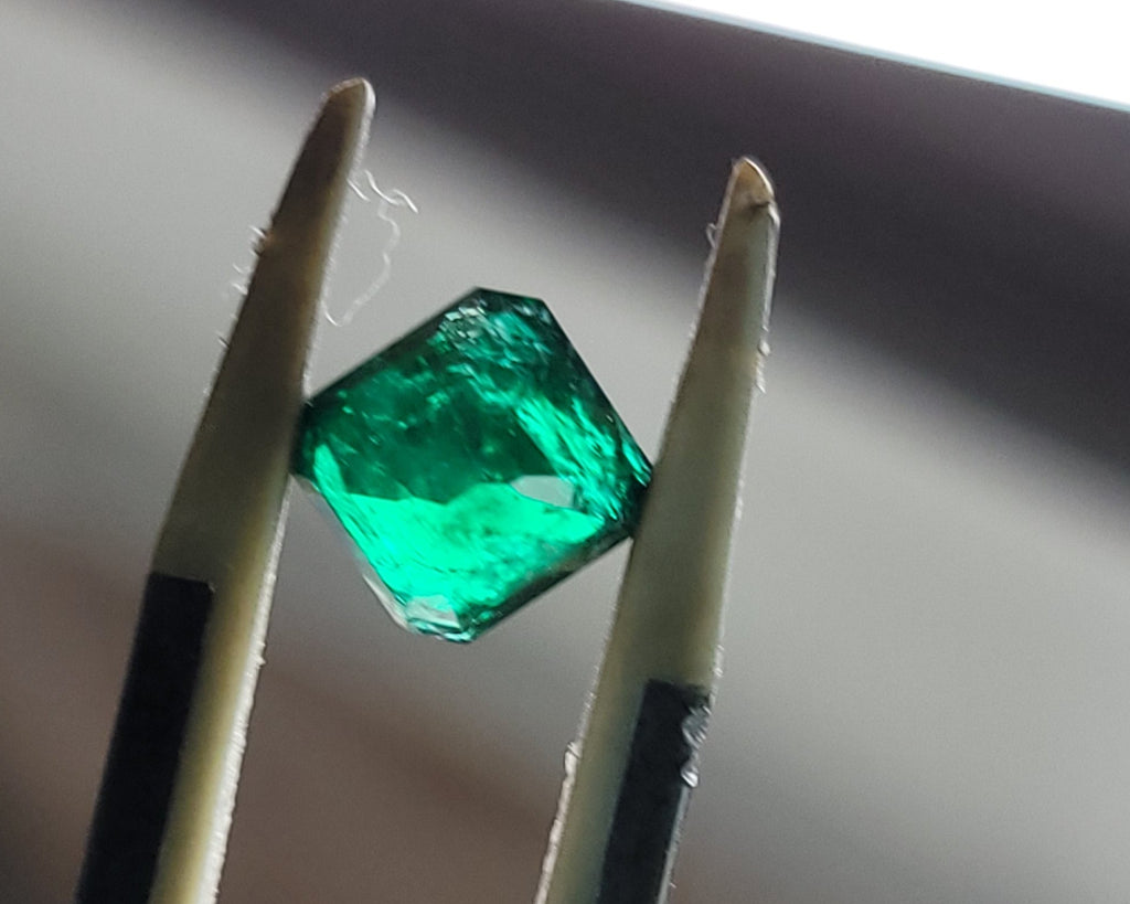 A faceted green gemstone held between two metal tweezers against a blurred gray background.