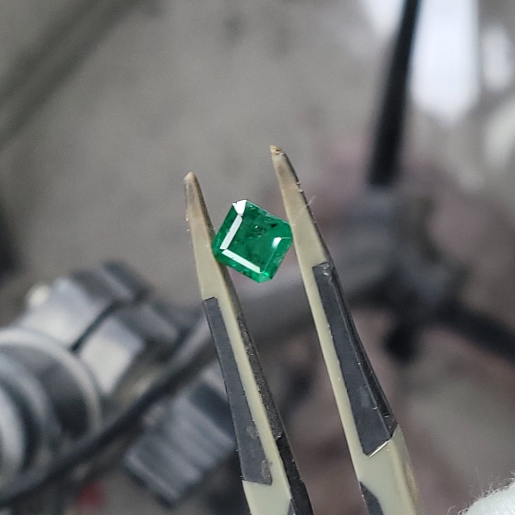 A small green faceted gemstone held between metal tweezers, with a blurred workshop or studio background.