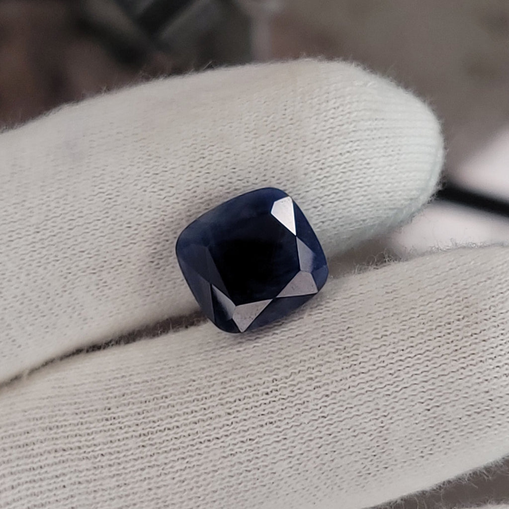 A loose cushion-cut dark blue gemstone held between white gloved fingers, with a blurred workshop or studio background.
