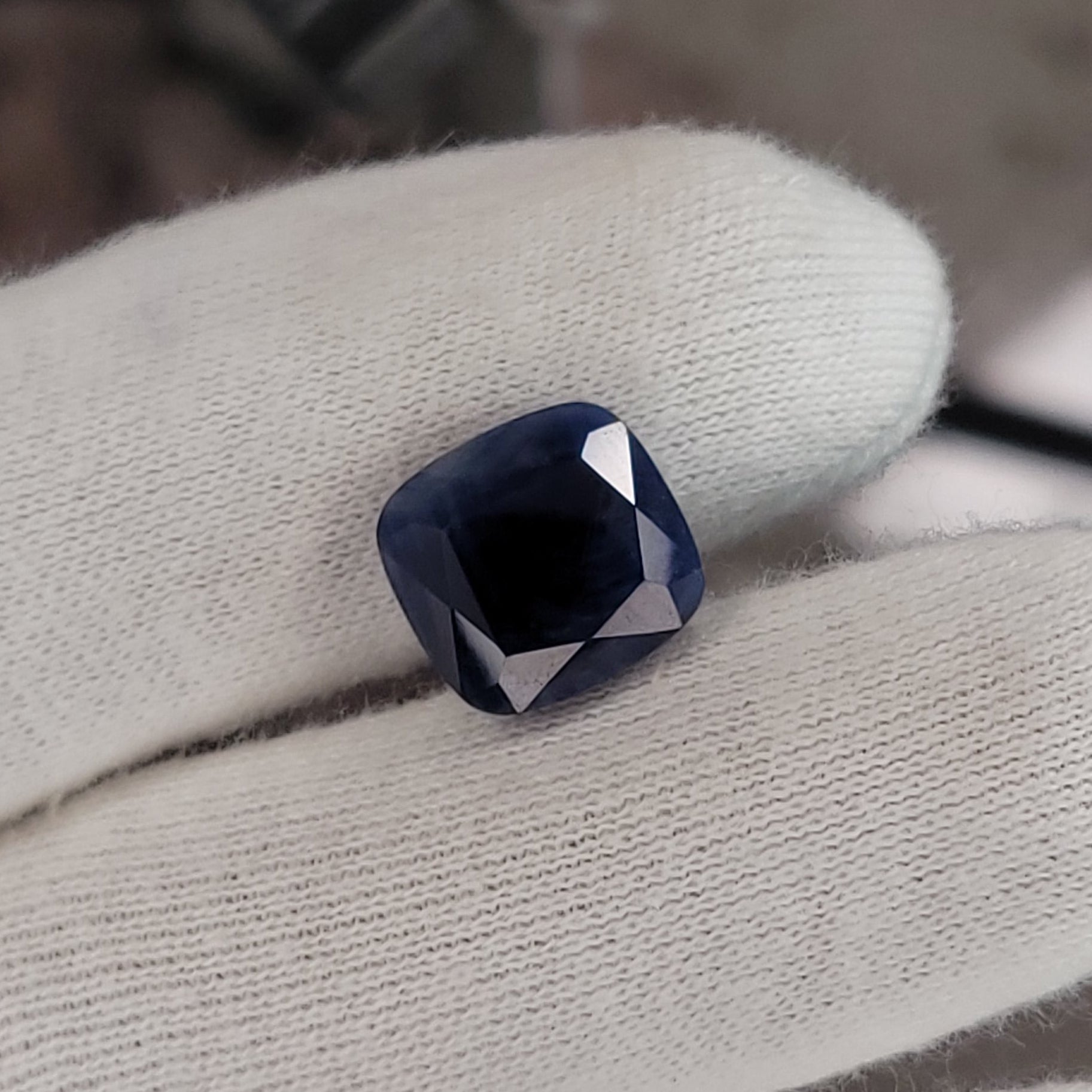 A loose cushion-cut dark blue gemstone held between white gloved fingers, with a blurred workshop or studio background.