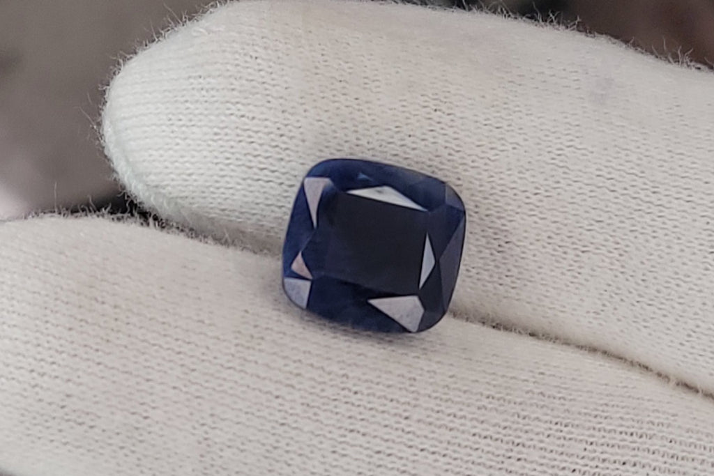 A cushion-cut deep blue gemstone held on a gloved finger, with a blurred studio or workshop background.