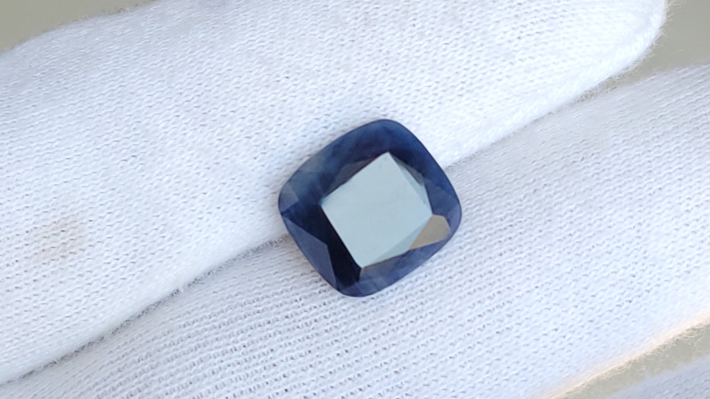 A square cushion-cut deep blue gemstone displayed on a white gloved finger.