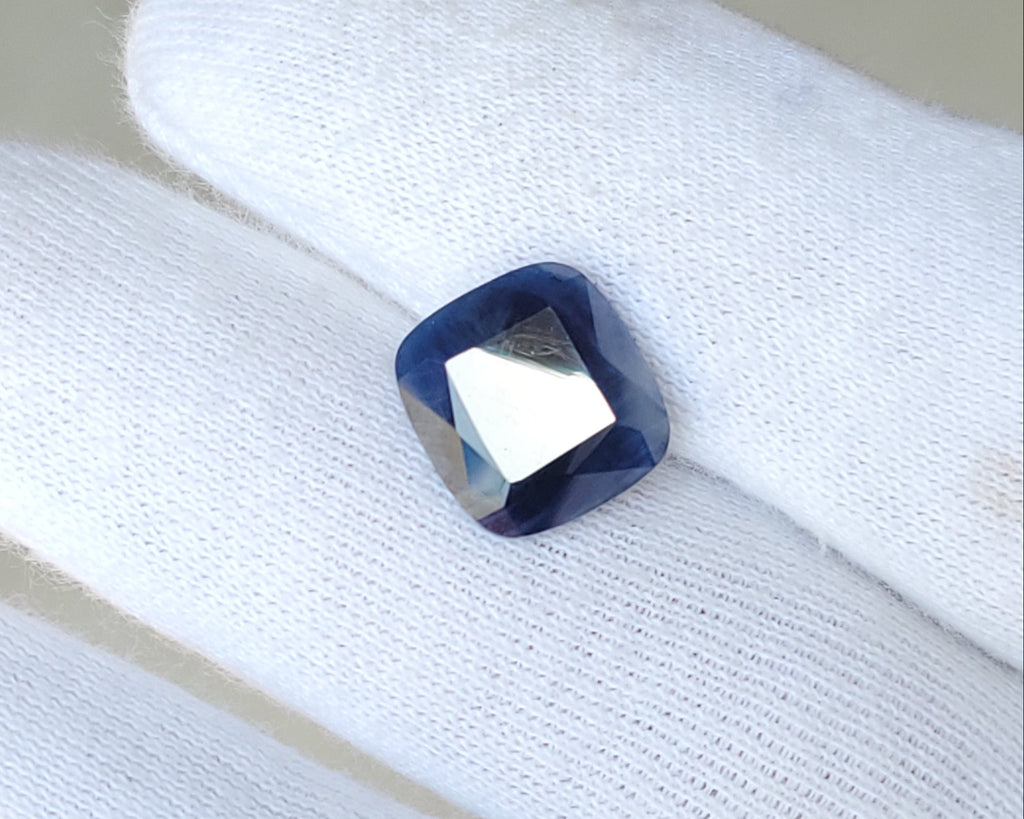 A small square-cut dark blue gemstone with a faceted center, resting on a white gloved finger.