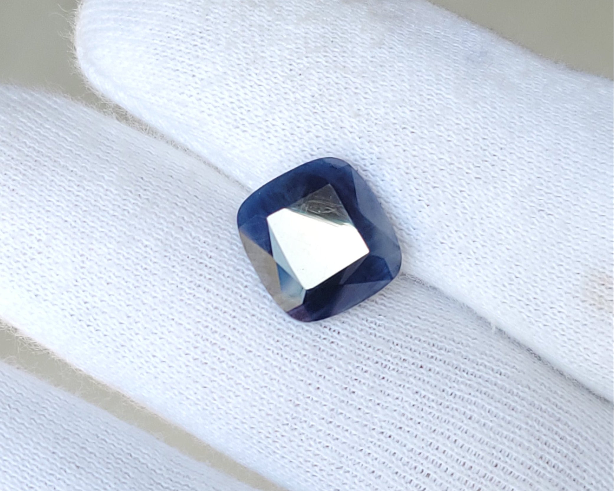 A small square-cut dark blue gemstone with a faceted center, resting on a white gloved finger.