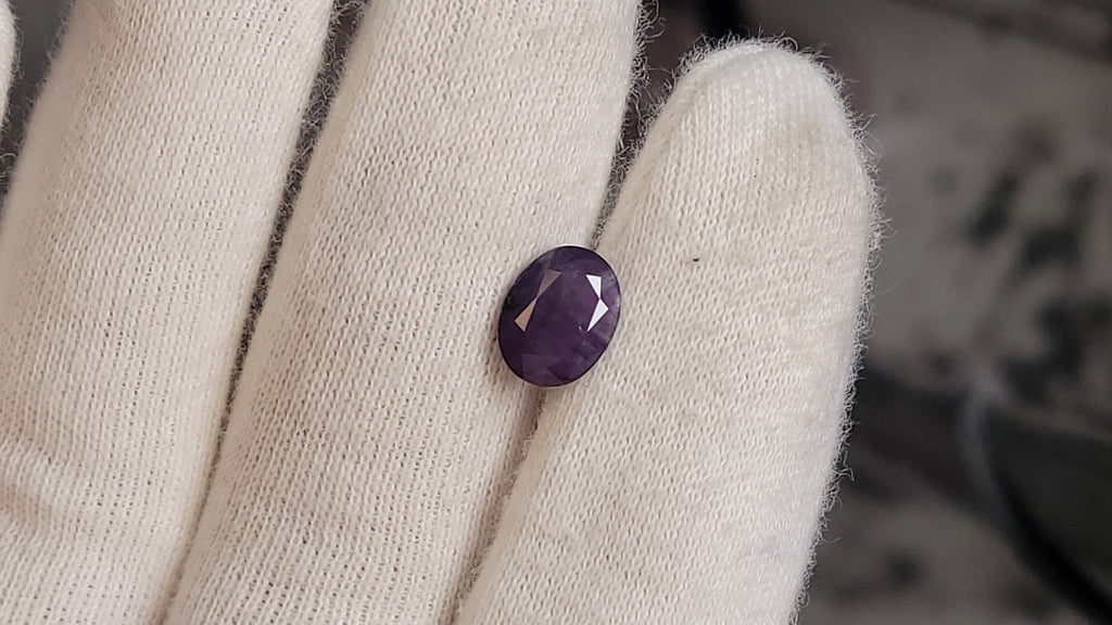 A small oval purple gemstone held on a white knit-gloved hand, with a dark blurred background.