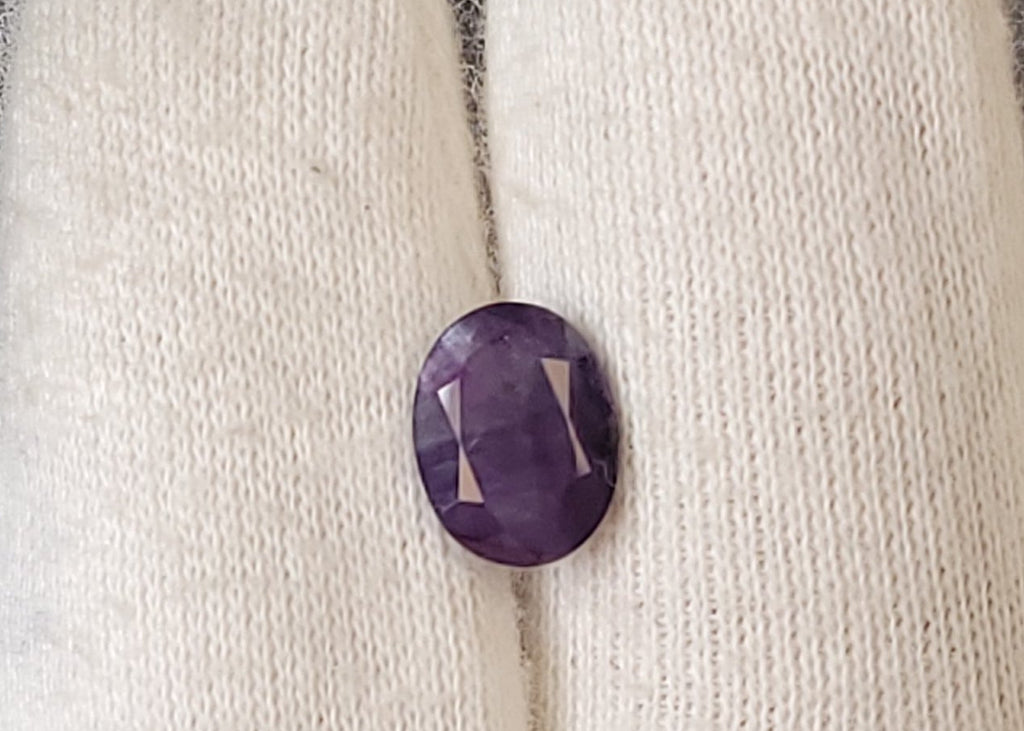 A small oval purple cabochon gemstone displayed on a white knit gloved finger.