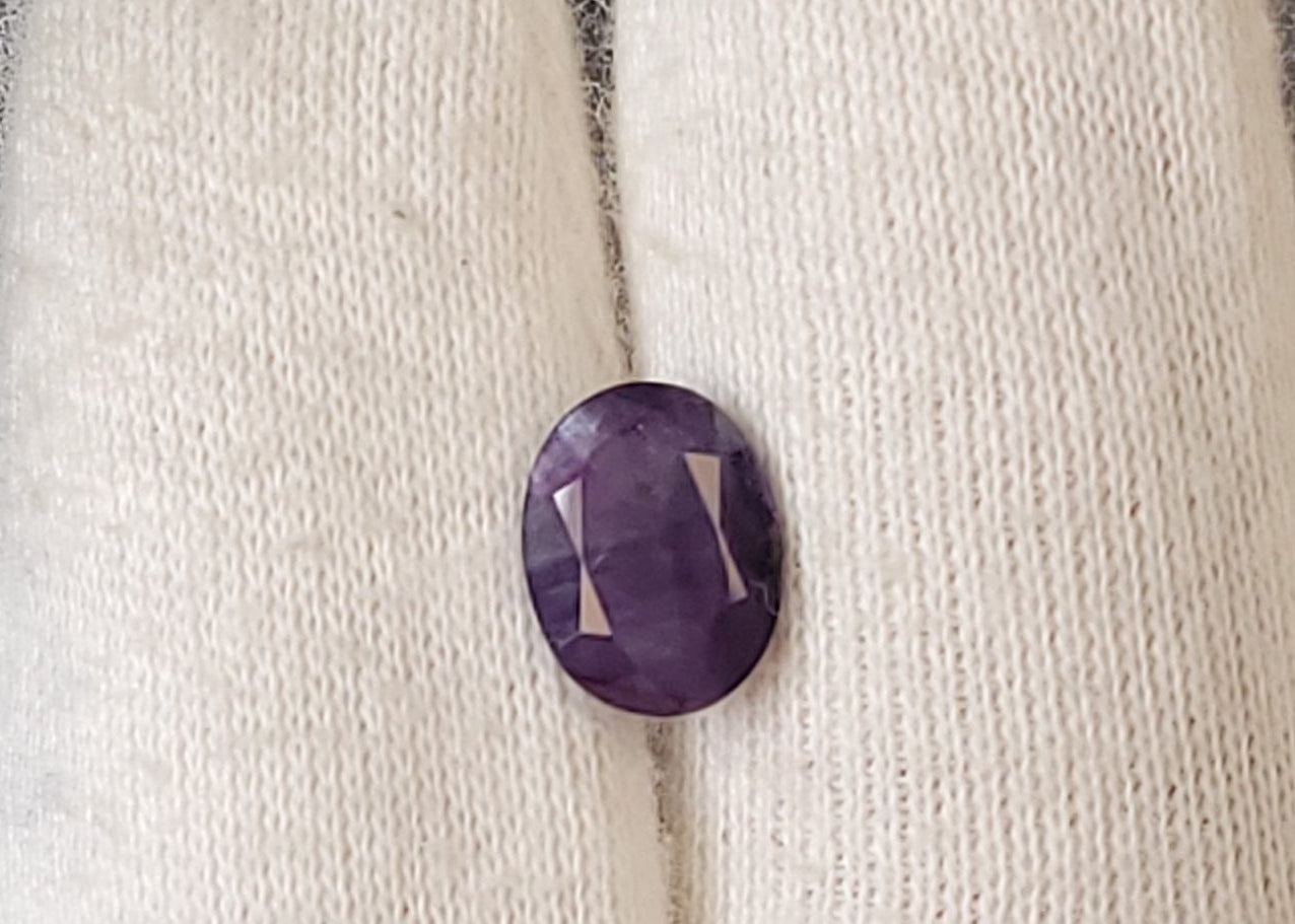 A small oval purple cabochon gemstone displayed on a white knit gloved finger.