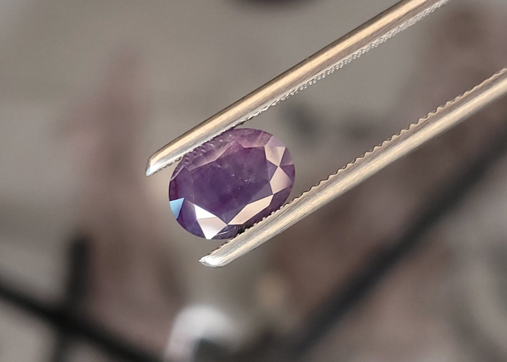 A faceted oval purple gemstone held by metal tweezers against a blurred workshop background.