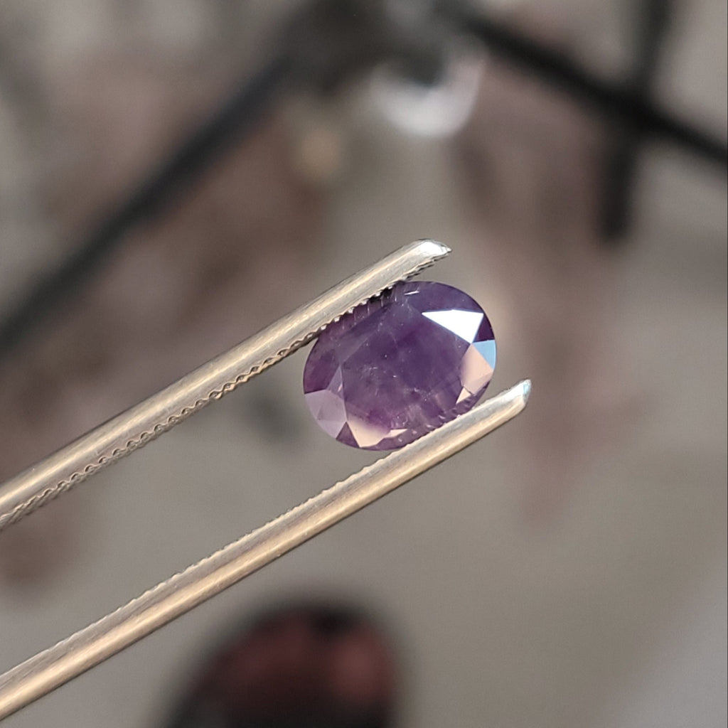 A faceted oval purple gemstone held in metal tweezers, shown close up against a blurred workshop background.