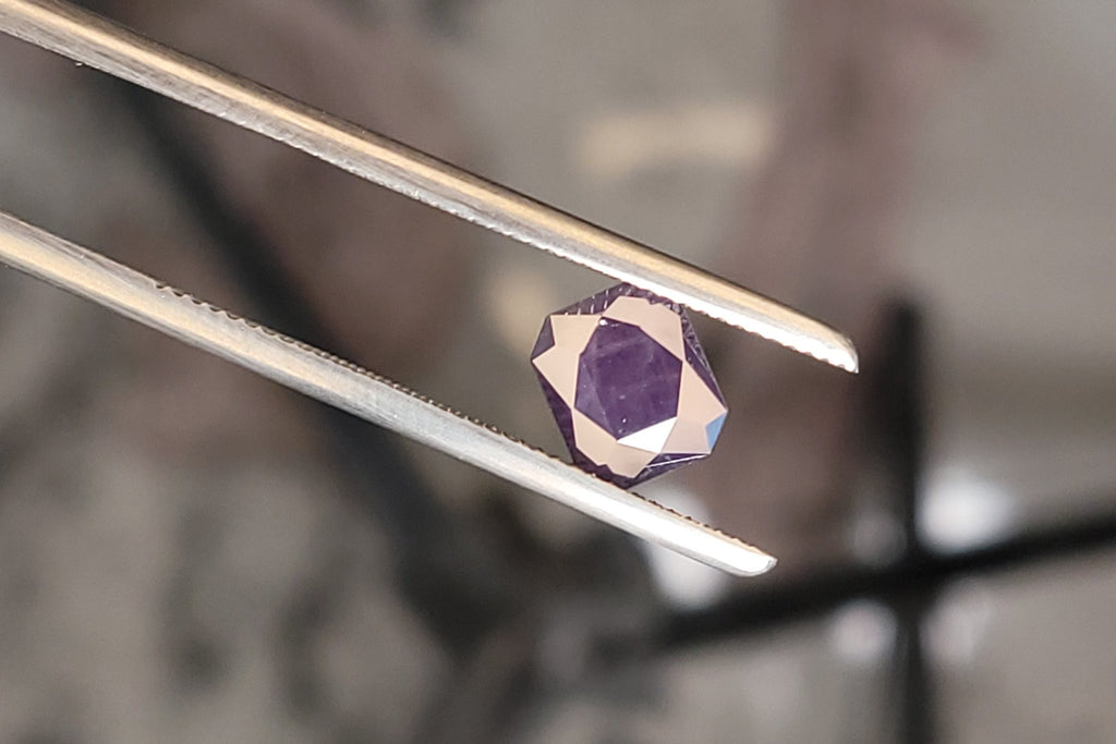 A faceted purple gemstone held between metal tweezers in a workshop setting with blurred tools and equipment in the background.