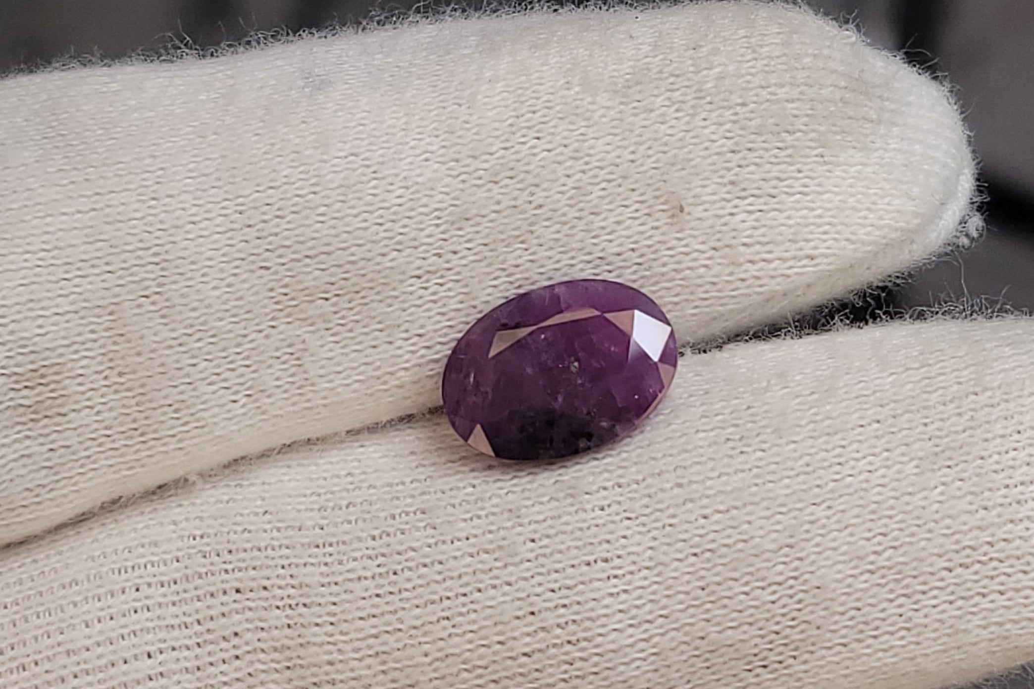 A small oval purple gemstone displayed on a white gloved finger, with a dark studio background and part of a camera visible at the left edge.
