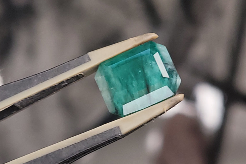 A green rectangular gemstone held in a pair of tweezers, shown in close-up against a blurred workshop background.