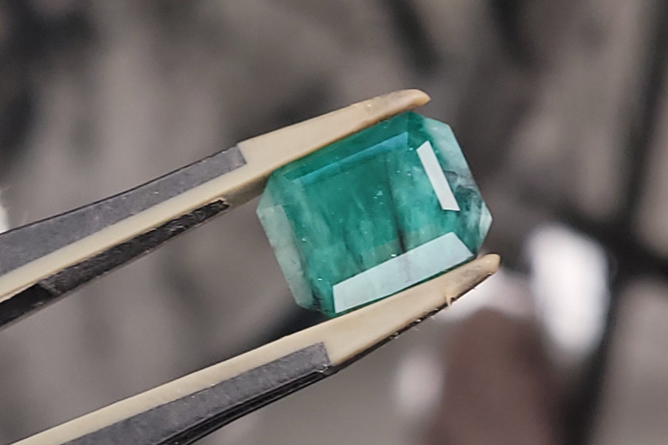 A green rectangular gemstone held in a pair of tweezers, shown in close-up against a blurred workshop background.