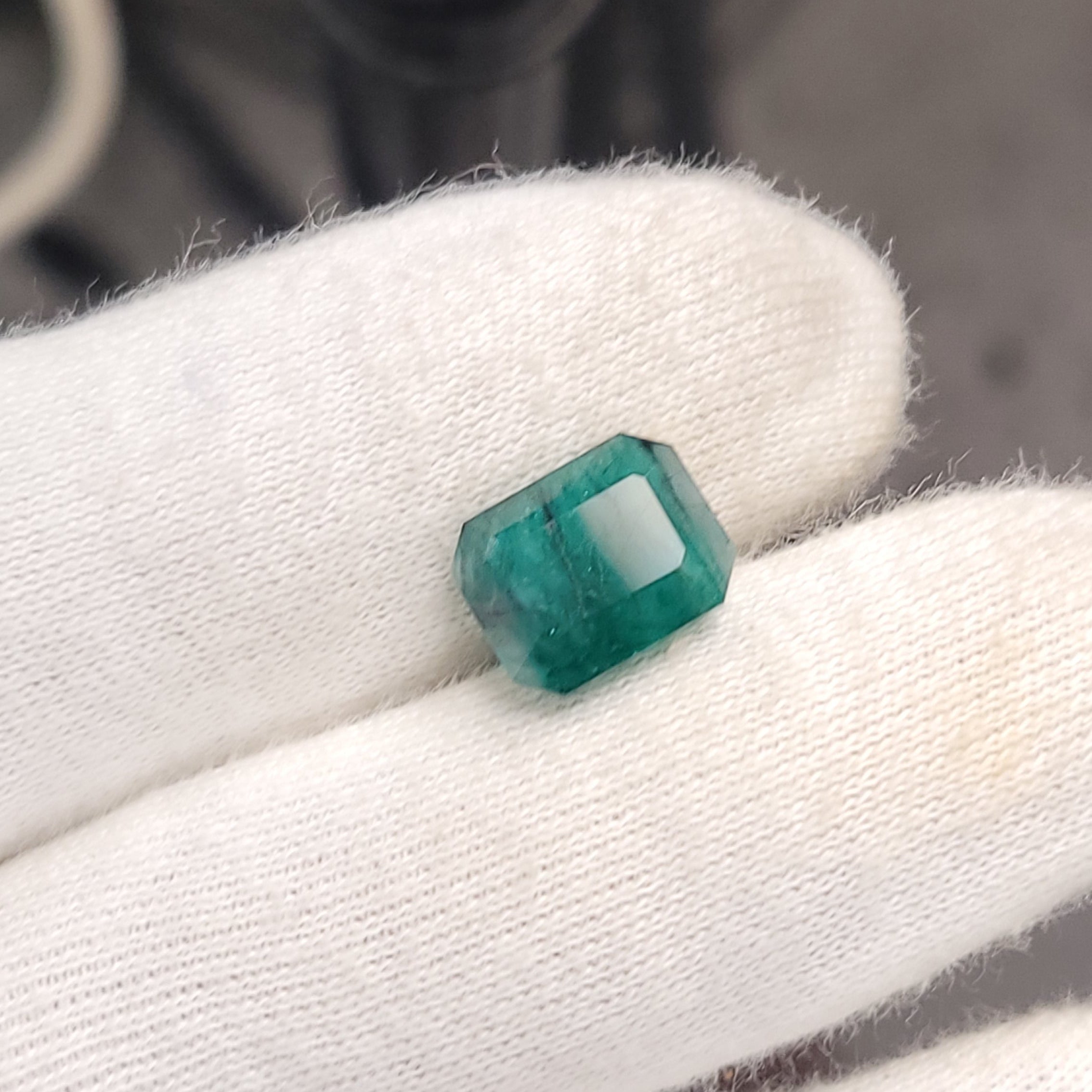 A small green rectangular gemstone held on a white gloved finger, with a blurred camera or tripod in the background.
