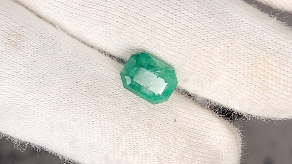 A small green faceted gemstone held on a white gloved hand.