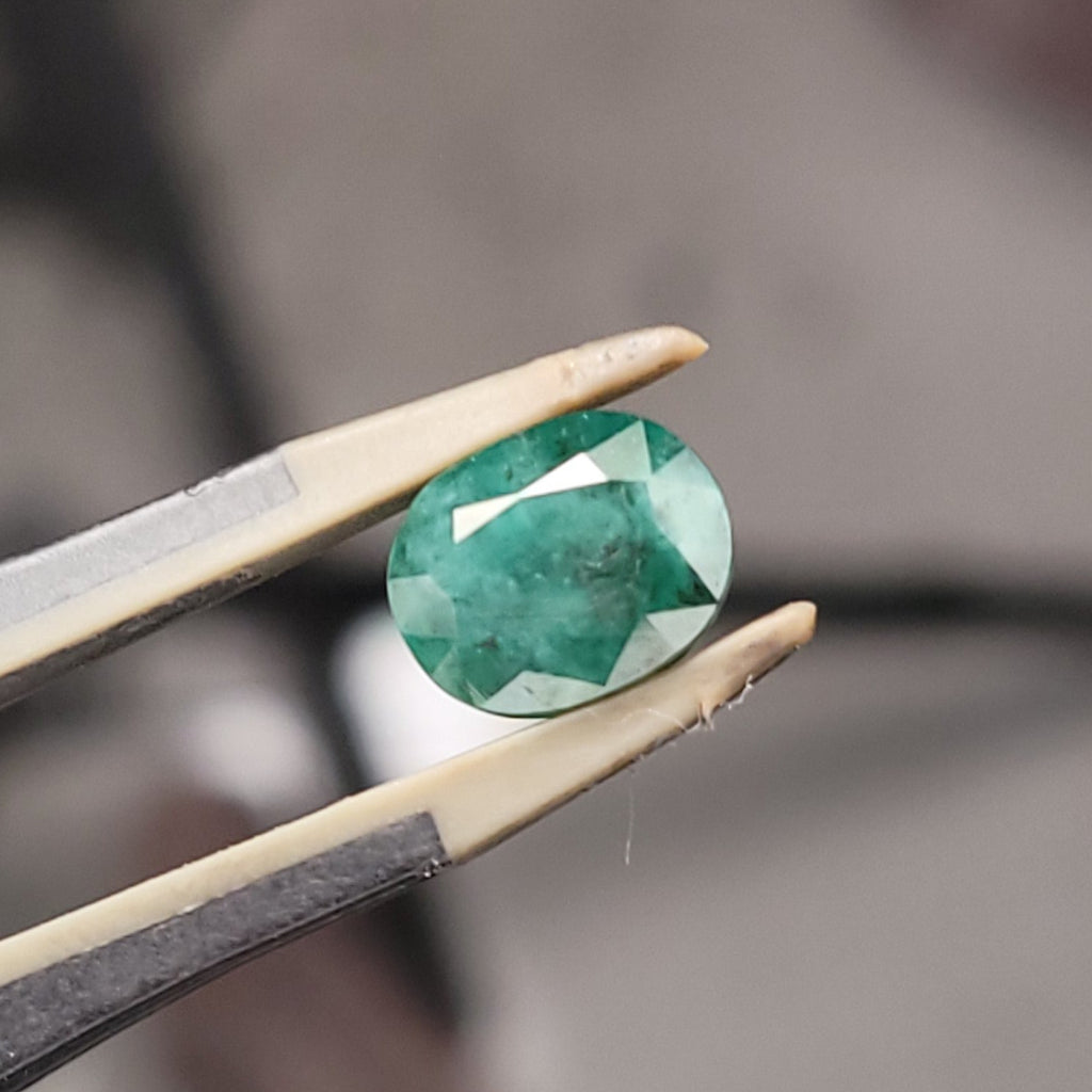 A green faceted gemstone held between metal tweezers against a blurred workshop background.