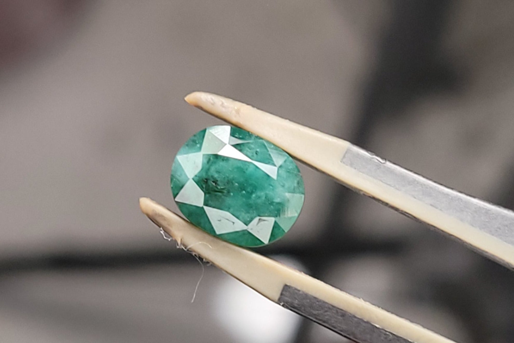 A faceted green gemstone held between metal tweezers against a blurred workshop background.