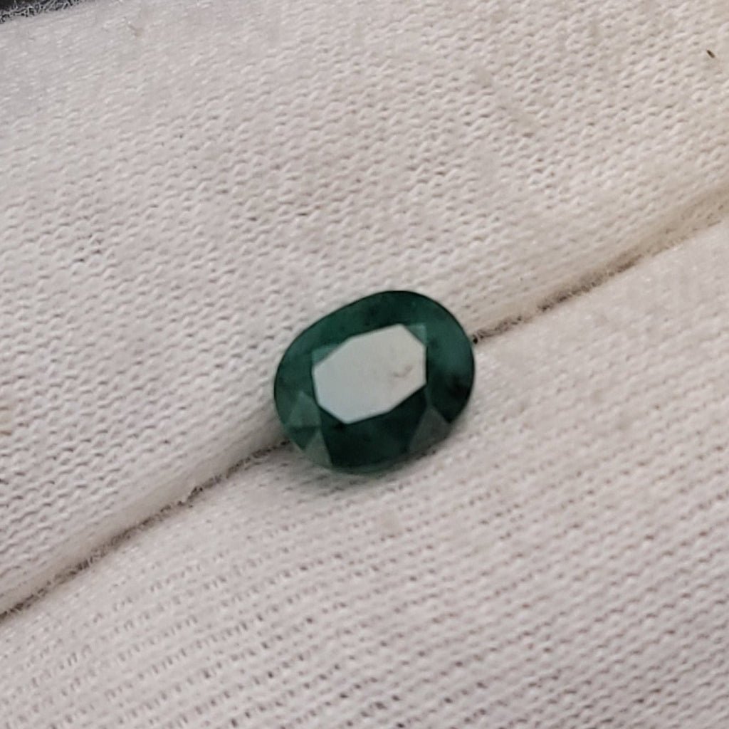 A small oval-cut dark green gemstone held on a white knit gloved finger, with a blurred dark background.