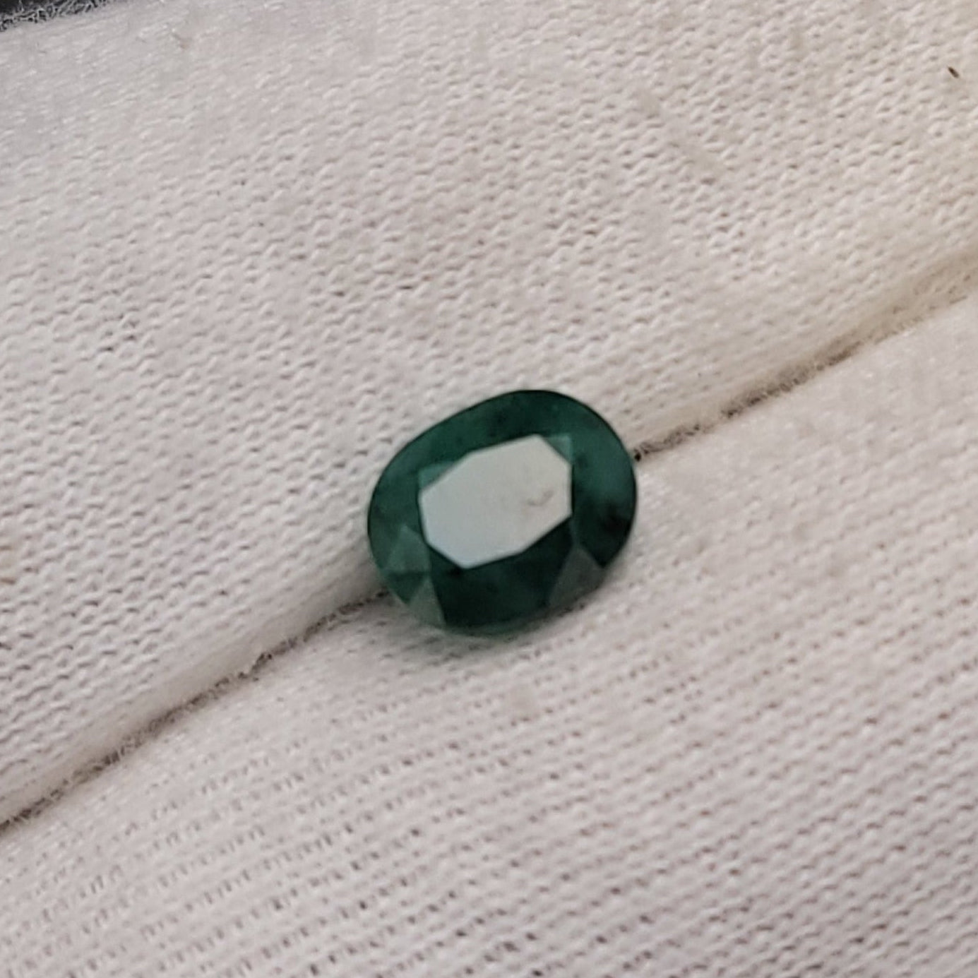 A small oval-cut dark green gemstone held on a white knit gloved finger, with a blurred dark background.