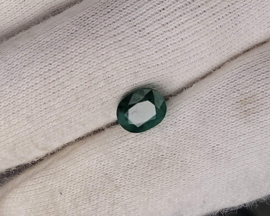 A small oval-cut dark green gemstone displayed on a white gloved hand.