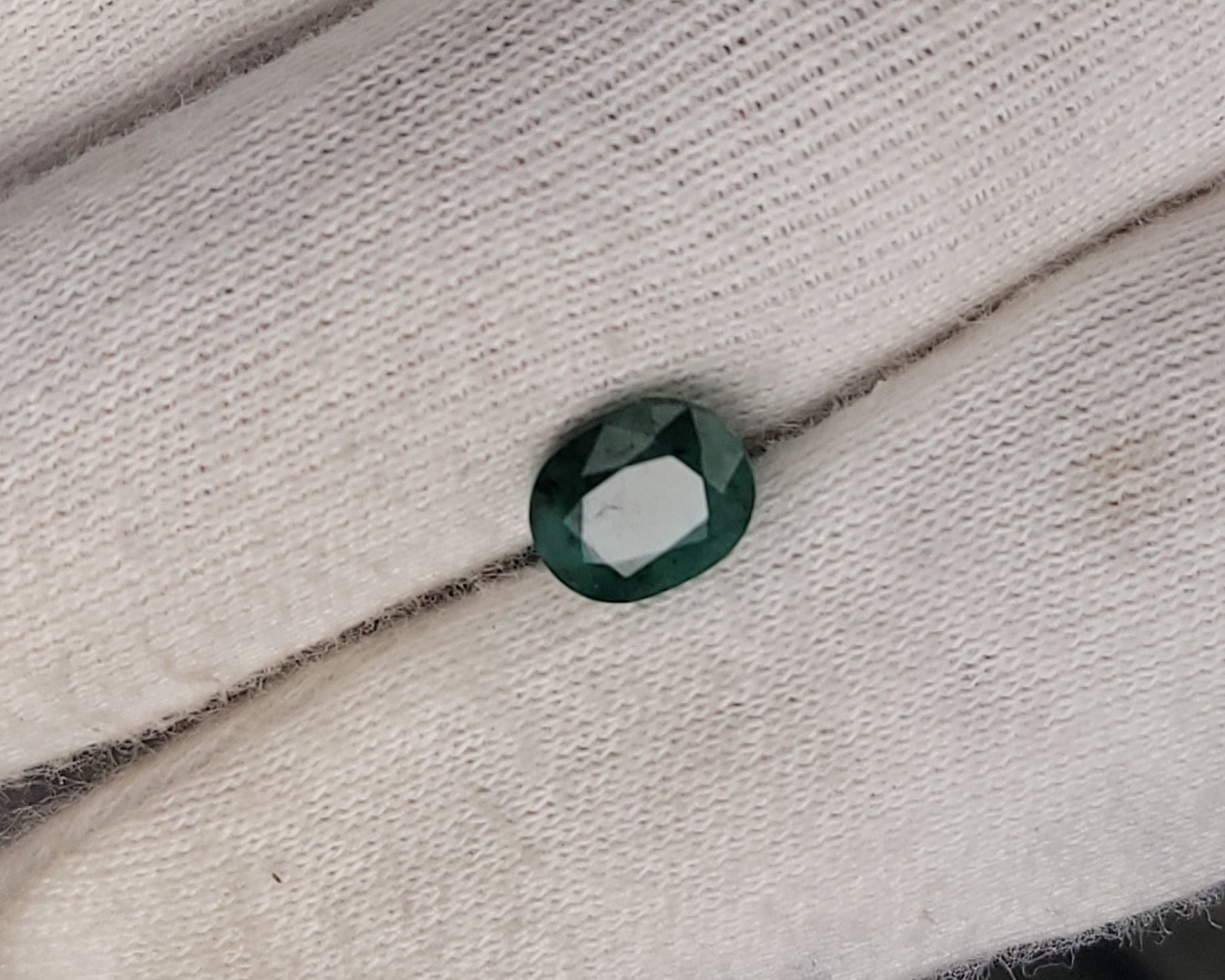 A small oval-cut dark green gemstone displayed on a white gloved hand.