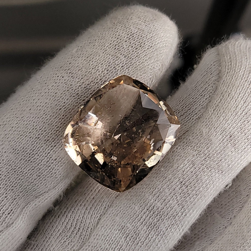 A cushion-cut, square-shaped champagne-colored gemstone held between white cotton gloves.