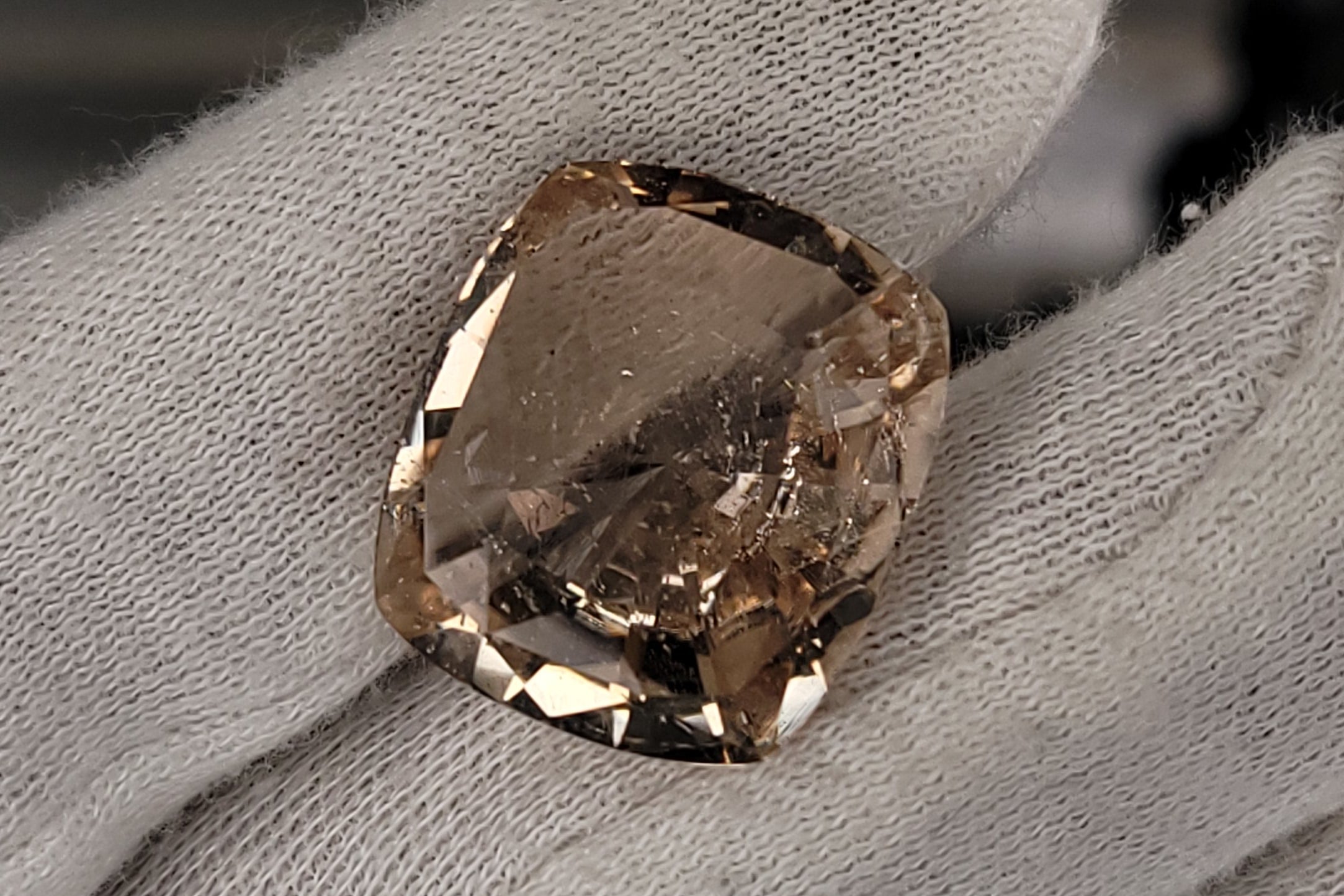 A rough, uncut cushion-cut gemstone with a champagne-gold and brown color identity, held between a white gloved fingers.