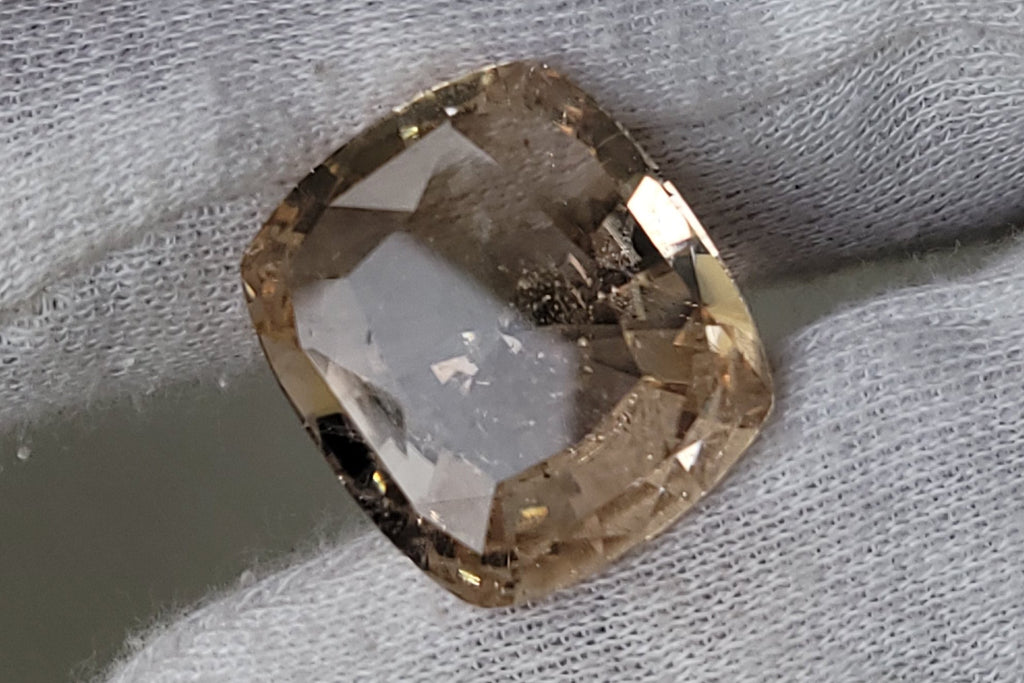 A cushion-cut, loose, pale champagne-colored gemstone held between a white fabric glove fingers against a neutral gray background.