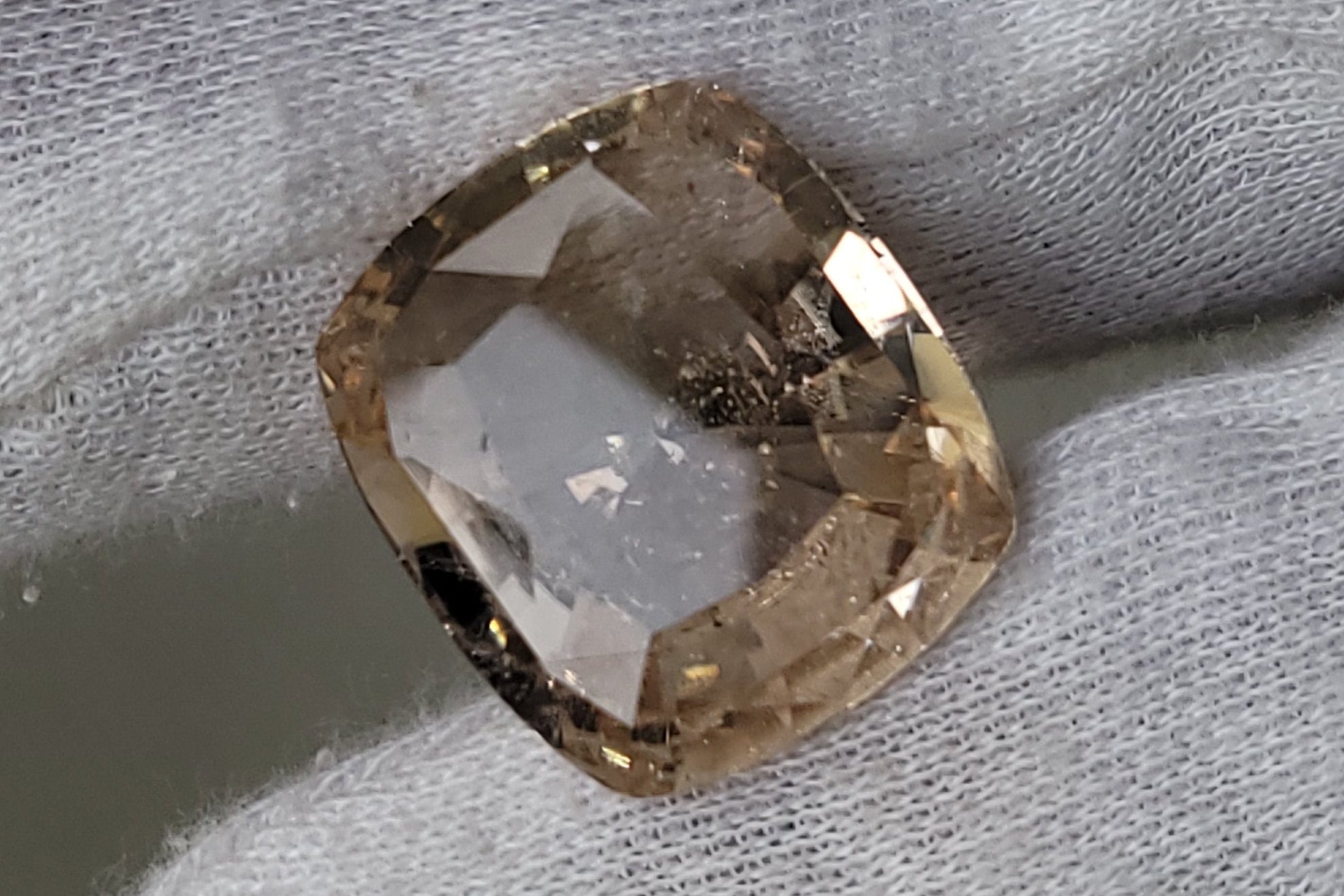 A cushion-cut, loose, pale champagne-colored gemstone held between a white fabric glove fingers against a neutral gray background.