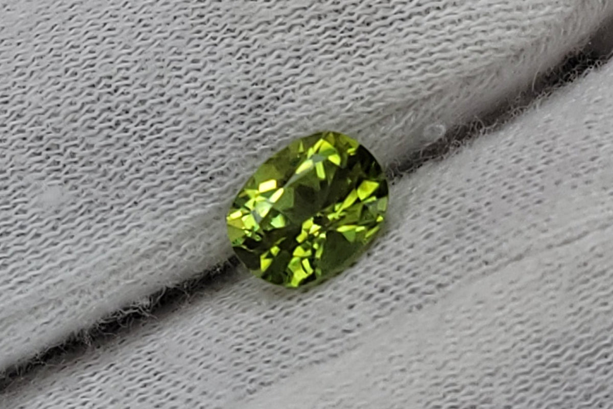 A small bright green gemstone, likely a cut peridot, held on the fingers of a hand wearing a white knit glove.