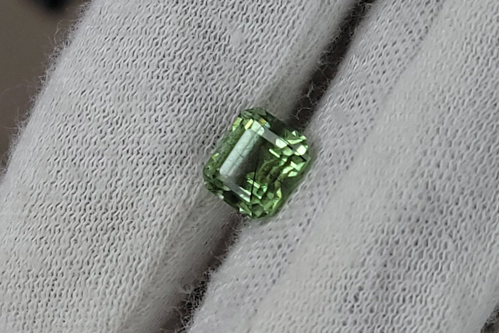 A small green rectangular gemstone held on a white textured glove, with a blurred indoor background.