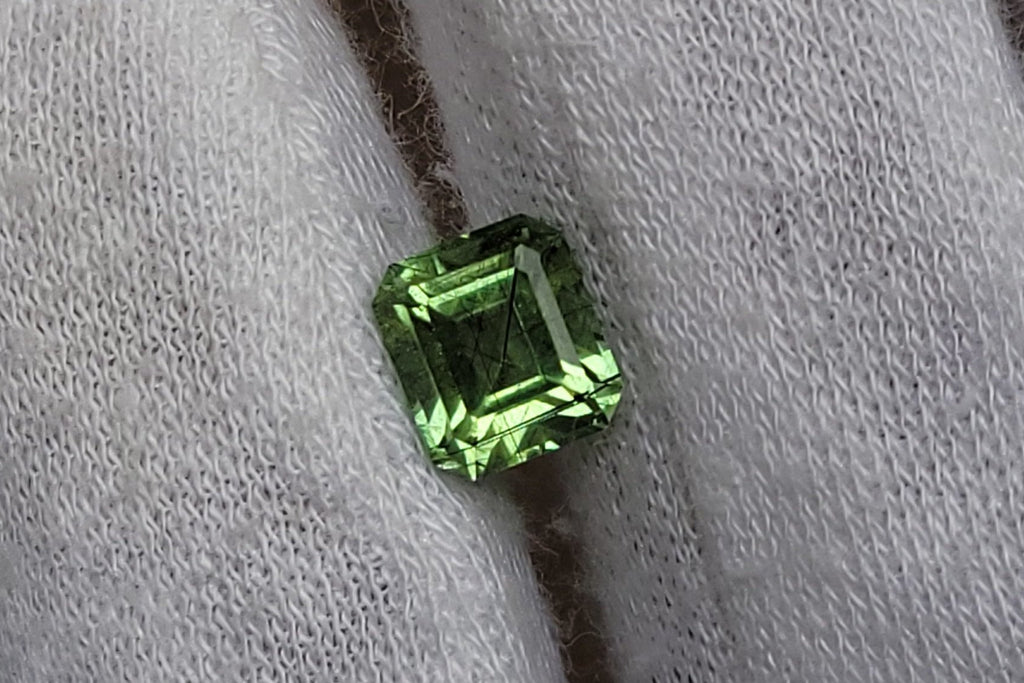 A small emerald-cut green gemstone displayed on a white knit gloved finger.