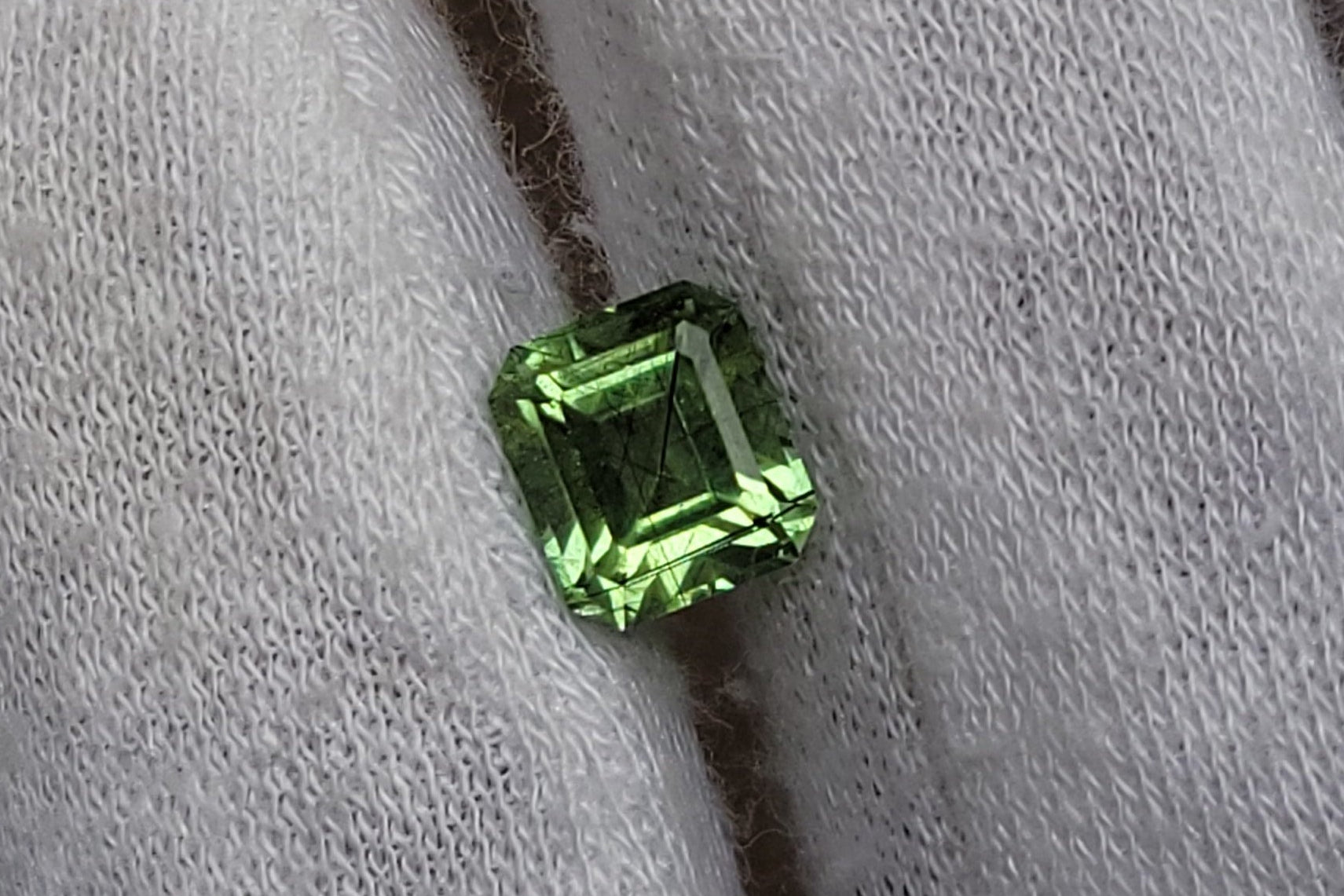 A small emerald-cut green gemstone displayed on a white knit gloved finger.