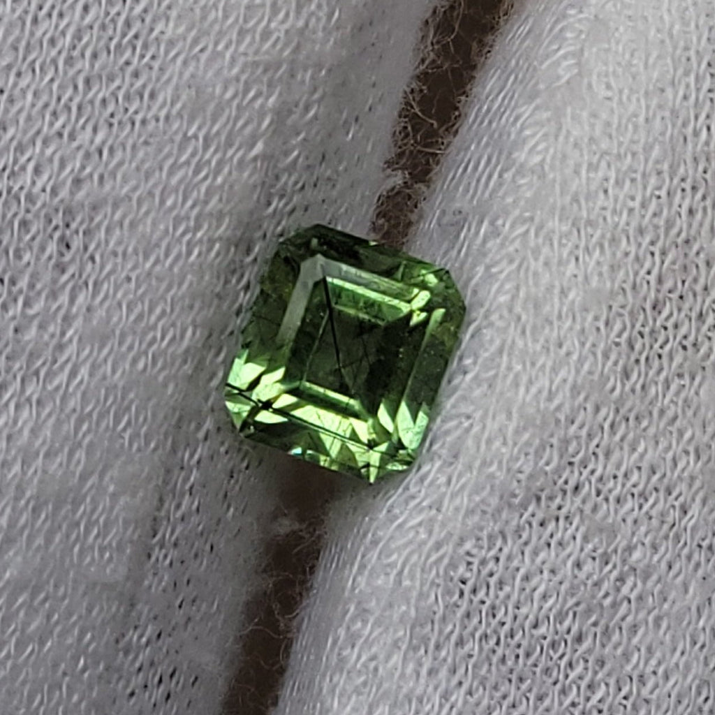 A small rectangular green gemstone held on a white gloved finger against a plain neutral background.