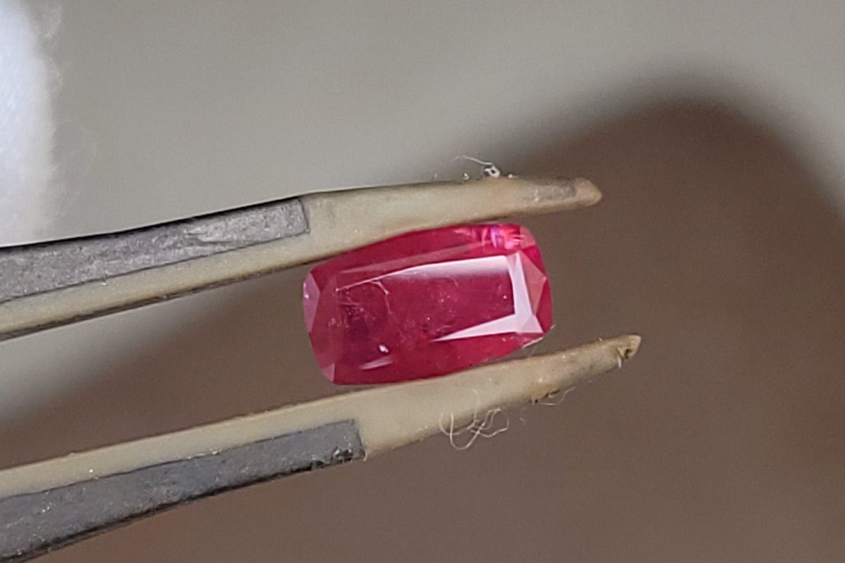 A small rectangular red gemstone held in metal tweezers between white gloved fingers, photographed close up against a soft gray and brown blurred background.