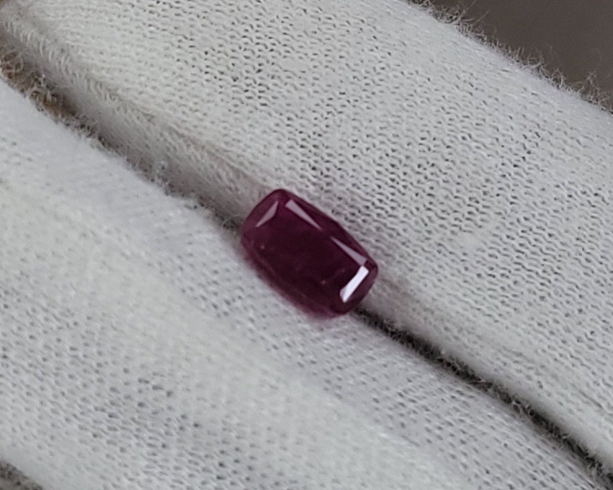 A small rectangular ruby gemstone resting on a white cotton glove, with a brown cardboard background.