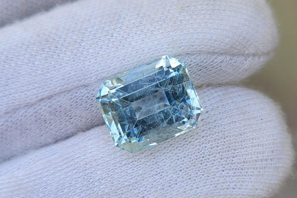 A rectangular light blue gemstone held on a gloved hand against a soft beige background.