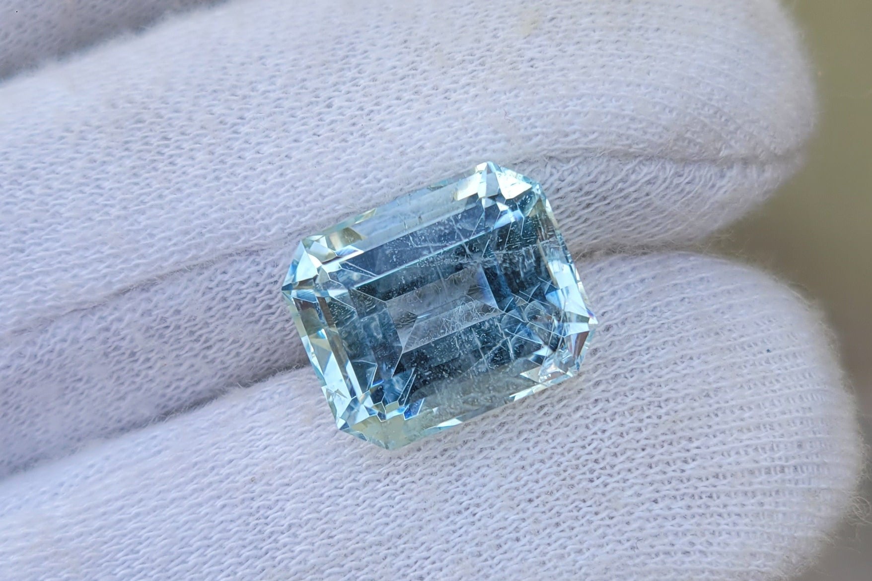 A rectangular light blue gemstone held on a gloved hand against a soft beige background.