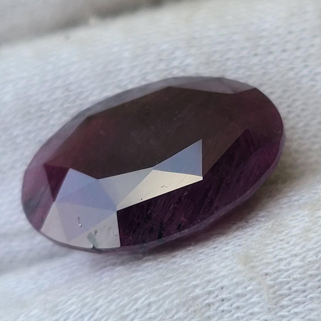 A faceted oval gemstone with a deep purple color, resting on a textured white fabric background.