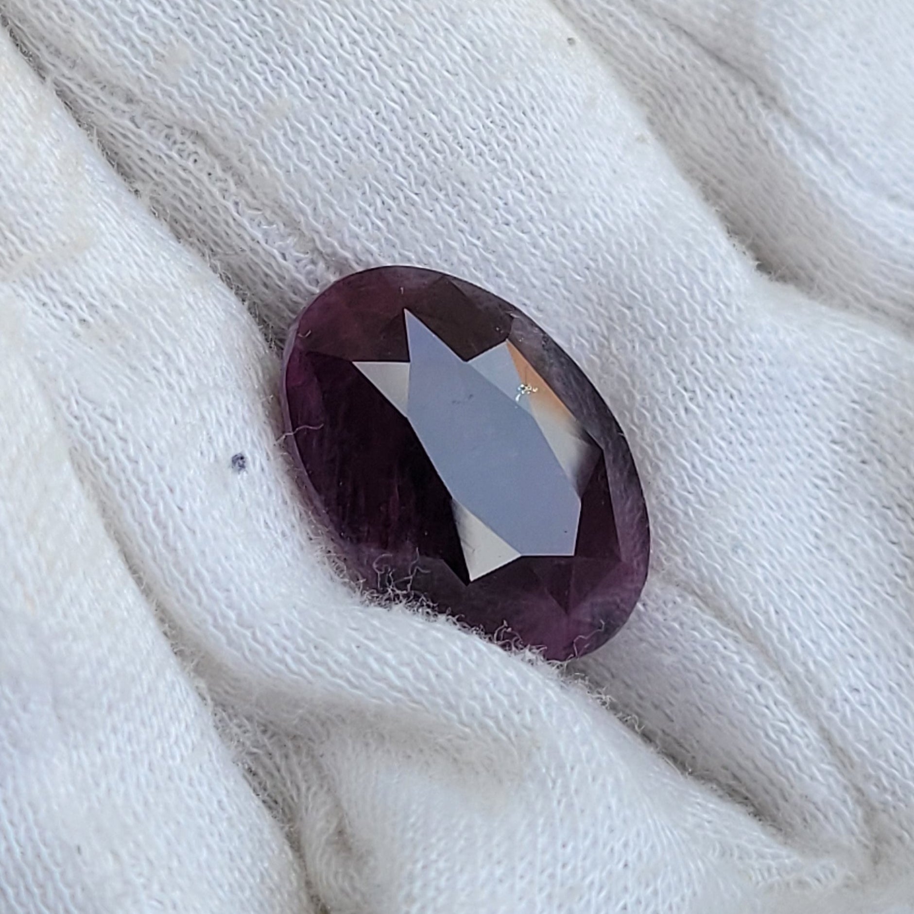 A small oval faceted dark purple gemstone resting on a white textured fabric surface.